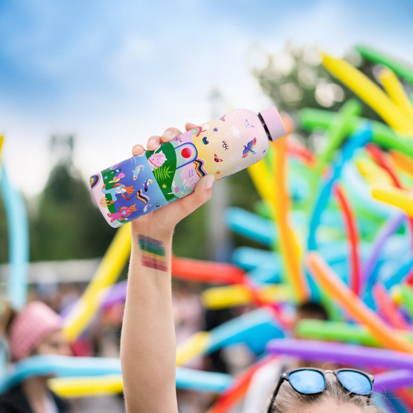 Badgie Be Yourself Steel Water Bottle BPAFree. Product image of a person holding the bottle in the air at a gay pride march LGBTQIA lgbt march event Mardi Gras parade, lesbian gay queer event with rainbows and colourful background.