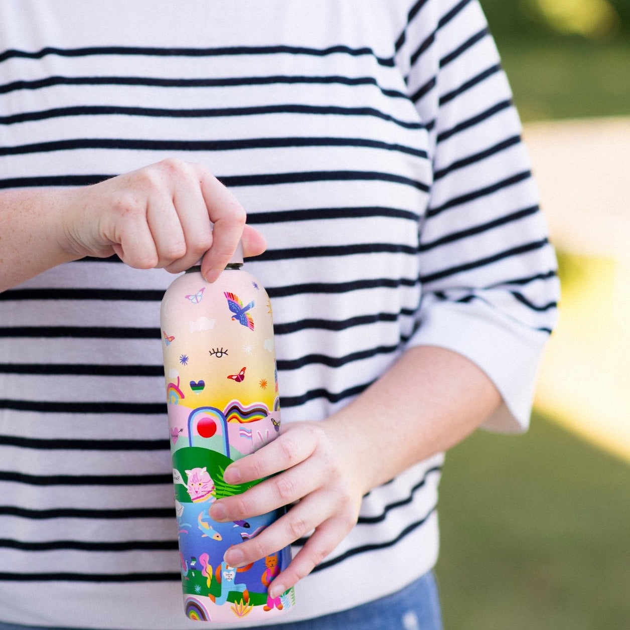 Badgie Be Yourself Steel Water Bottle BPAFree. Product image of a person holding the steel bottle bpafree, opening the lid, showing the colourful rainbow design with Australian artistry