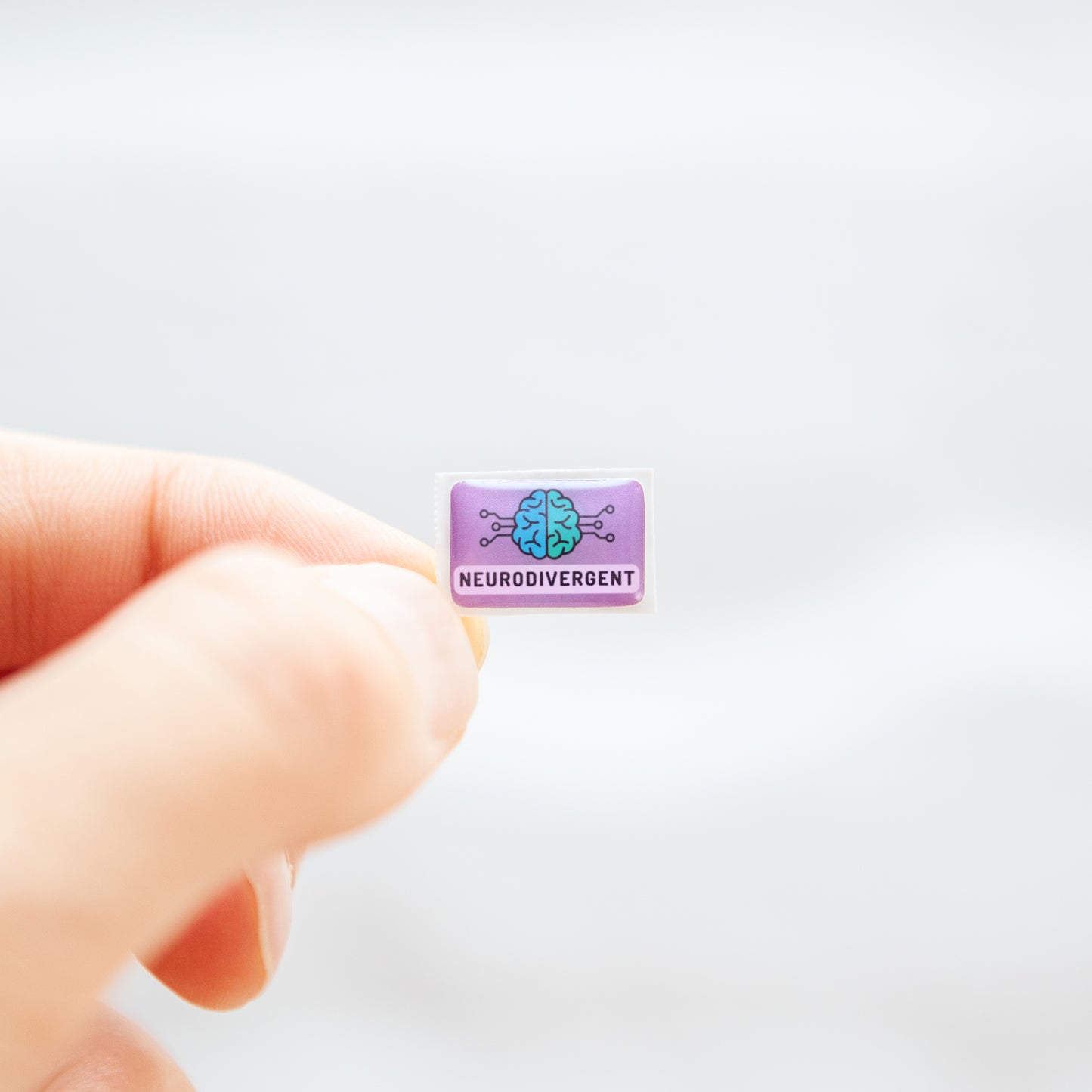 Fingers holding a lavender sticker, with 'Neurodivergent' text, and a digitally illustrated image of a brain, representing neurodivergent stickers.