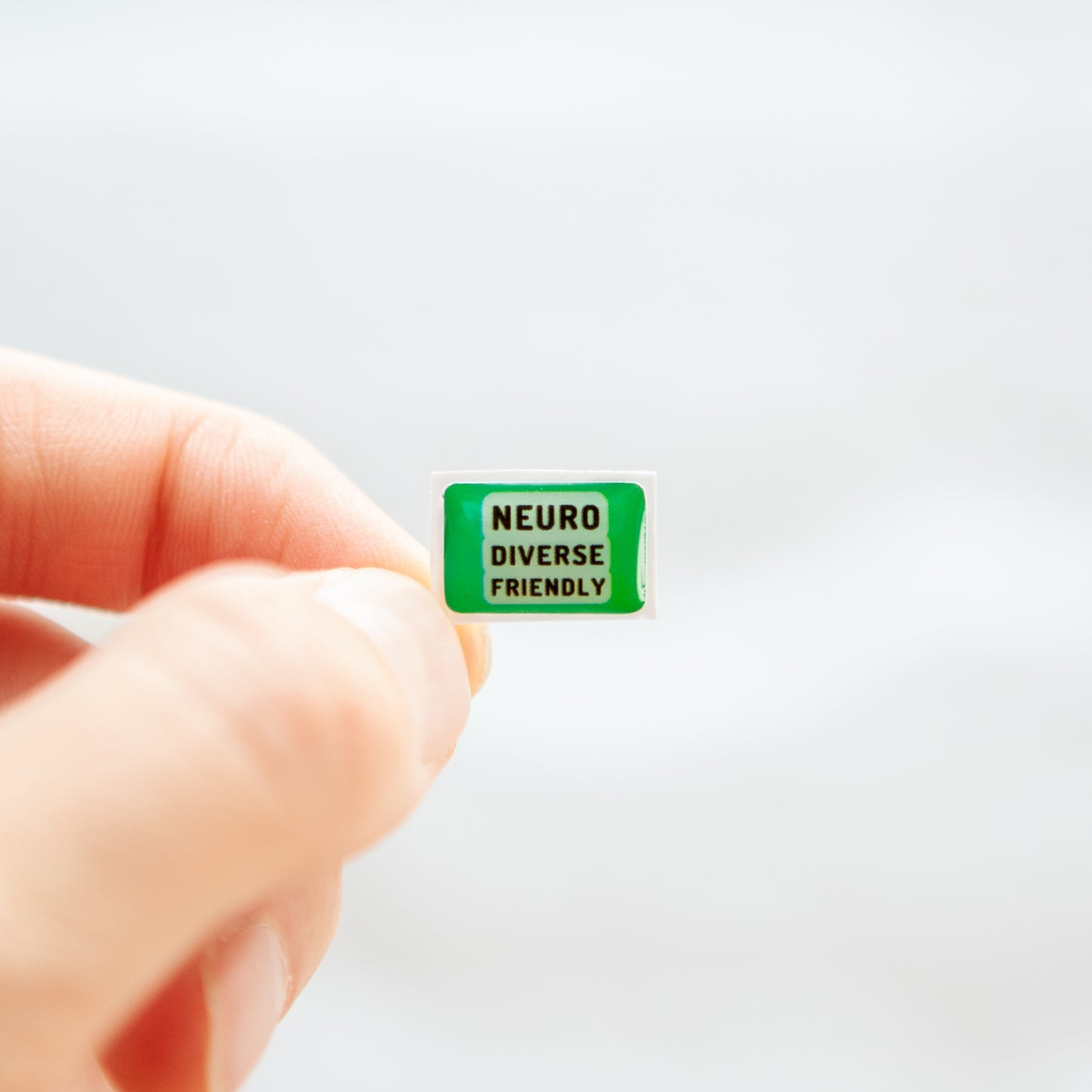 Fingers holding a green neurodivergent sticker with the text 'Neurodiverse Friendly'.