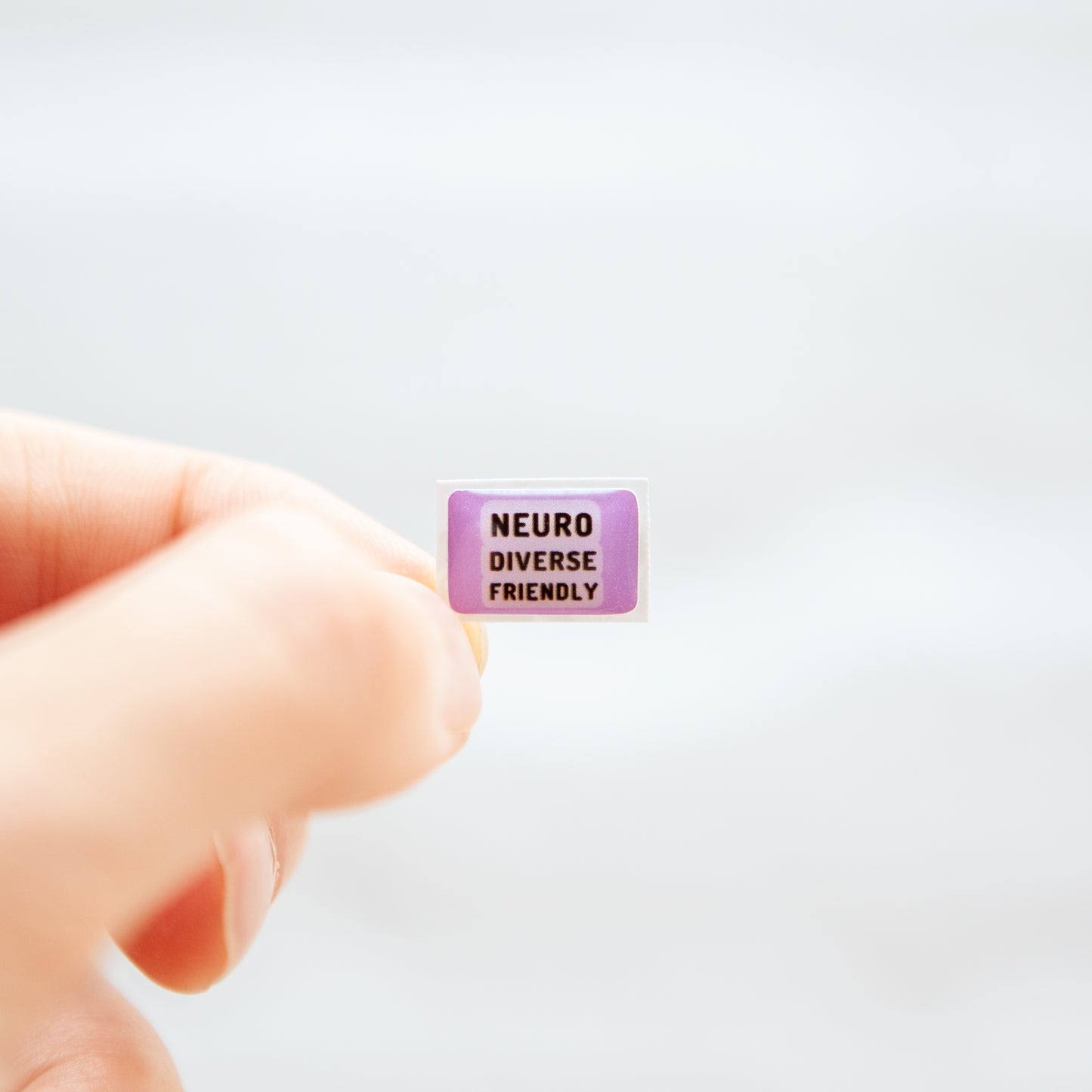 Fingers holding a lavender neurodivergent sticker that reads 'Neurodiverse Friendly'.