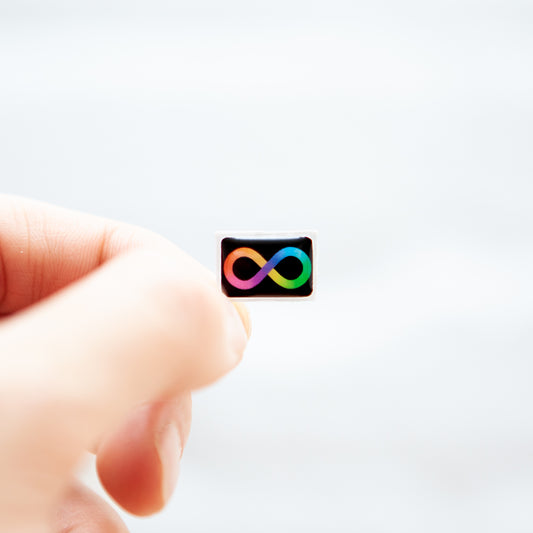 A hand holding a Badgie Neurodivergent Sticker, with a rainbow infinity loop design, showcasing the epoxy resin finish and high-quality details.