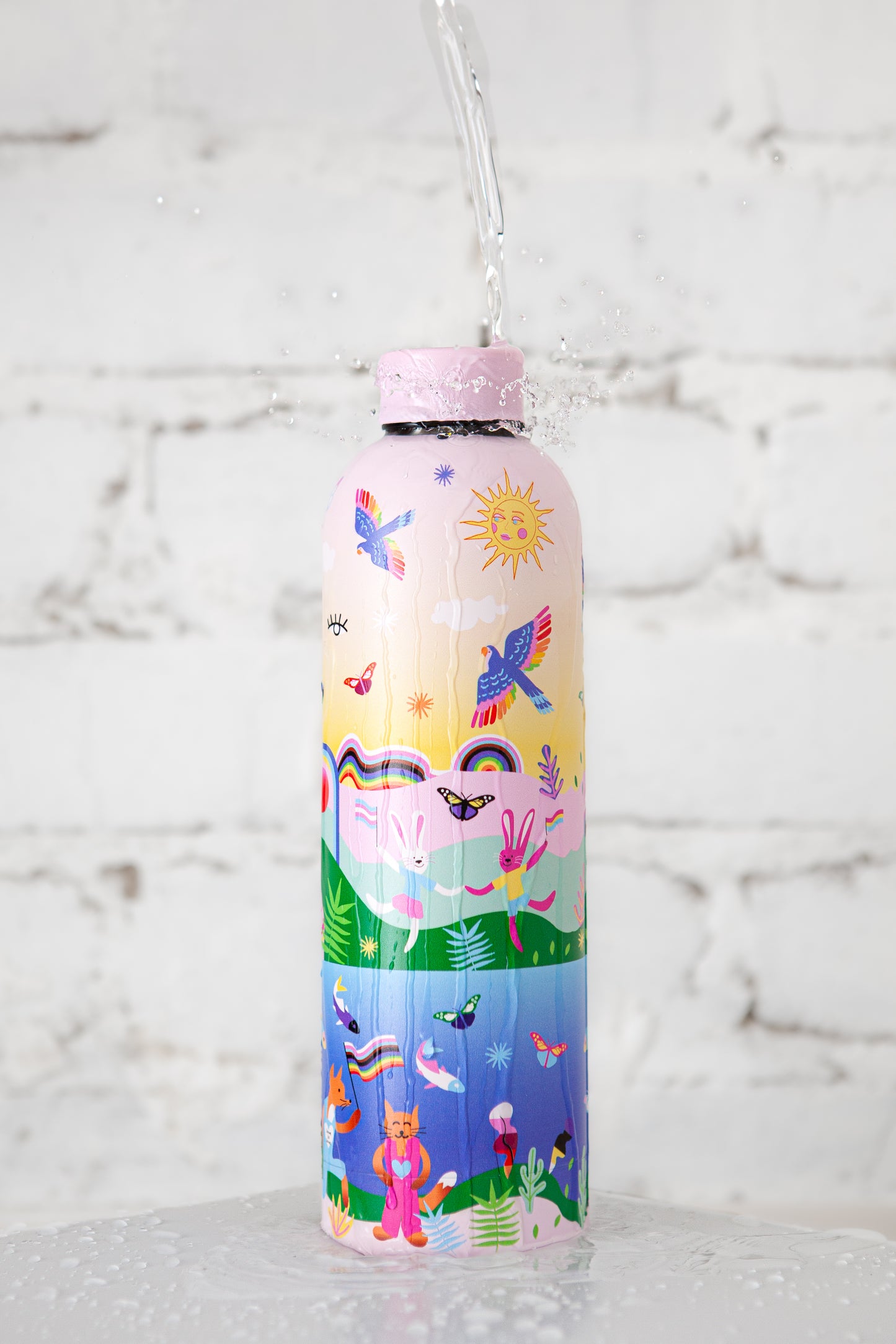 Badgie Be Yourself Steel Water Bottle BPAFree. Product image of steel bottle bpafree against white aesthetic brick background, with water pouring over the bottle, splashing around the colourful rainbow design with Australian artistry and lgbt pride gay lesbian queer colours