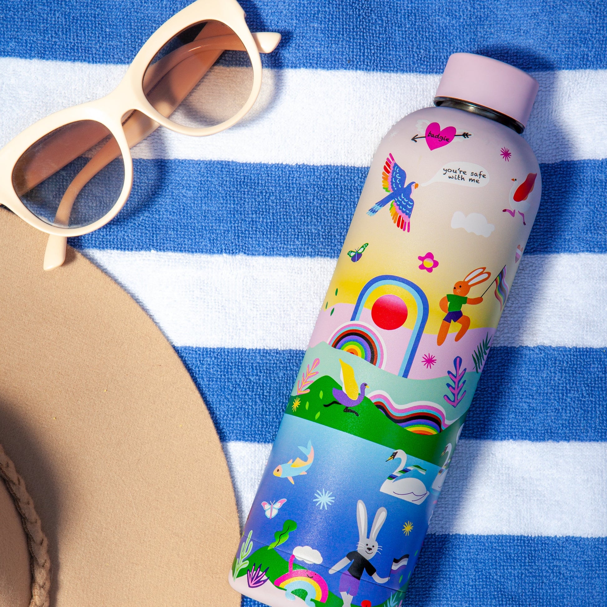 Badgie Be Yourself Steel Water Bottle BPAFree. Product image of the steel bottle at the beach, on a towel, used for travel and adventure, with sunglasses and hat visible