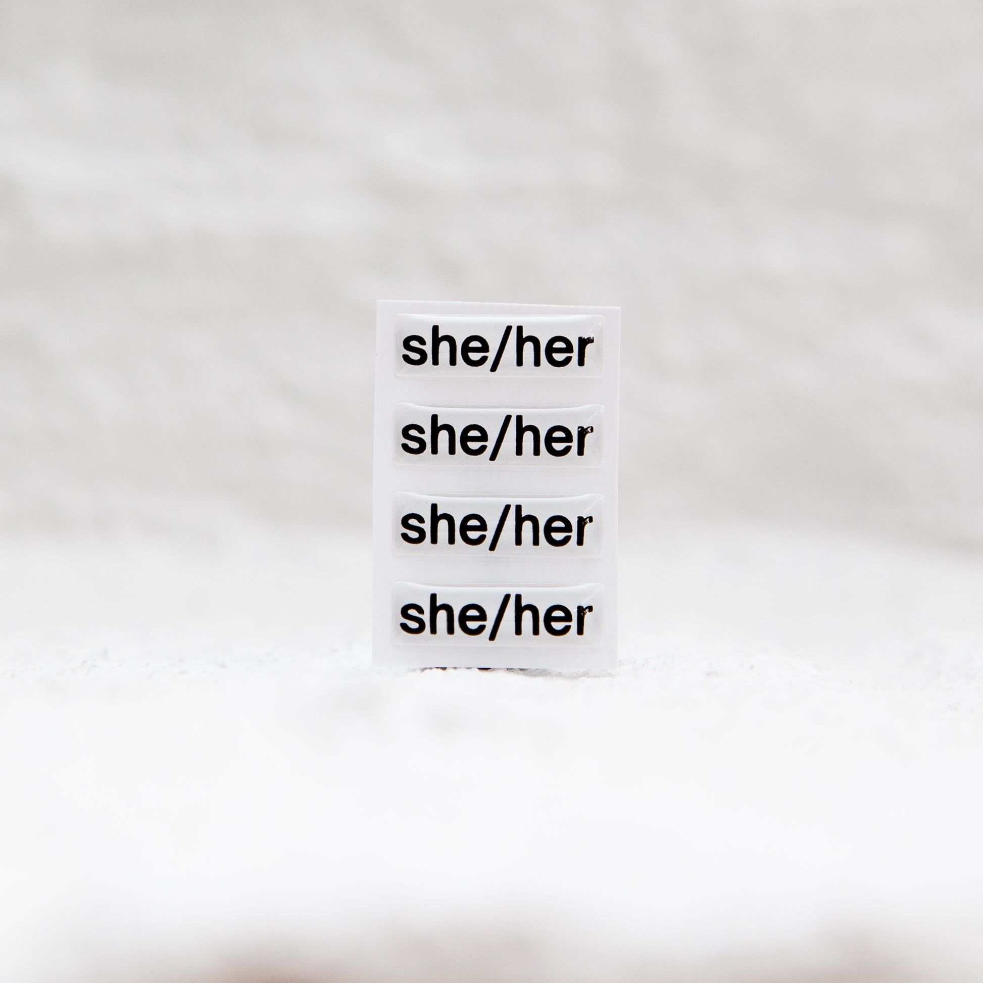 A white sticker with black text displaying she/her pronouns, designed for name badges and ID tags. A high-quality pronoun sticker with durable epoxy resin. This pronouns sticker for name badges