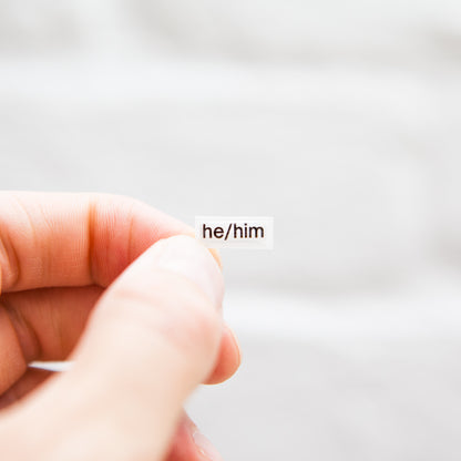 A white sticker with black text displaying he/him pronouns, designed for name badges and ID tags. A high-quality pronoun sticker with durable epoxy resin. This pronouns sticker for name badges.