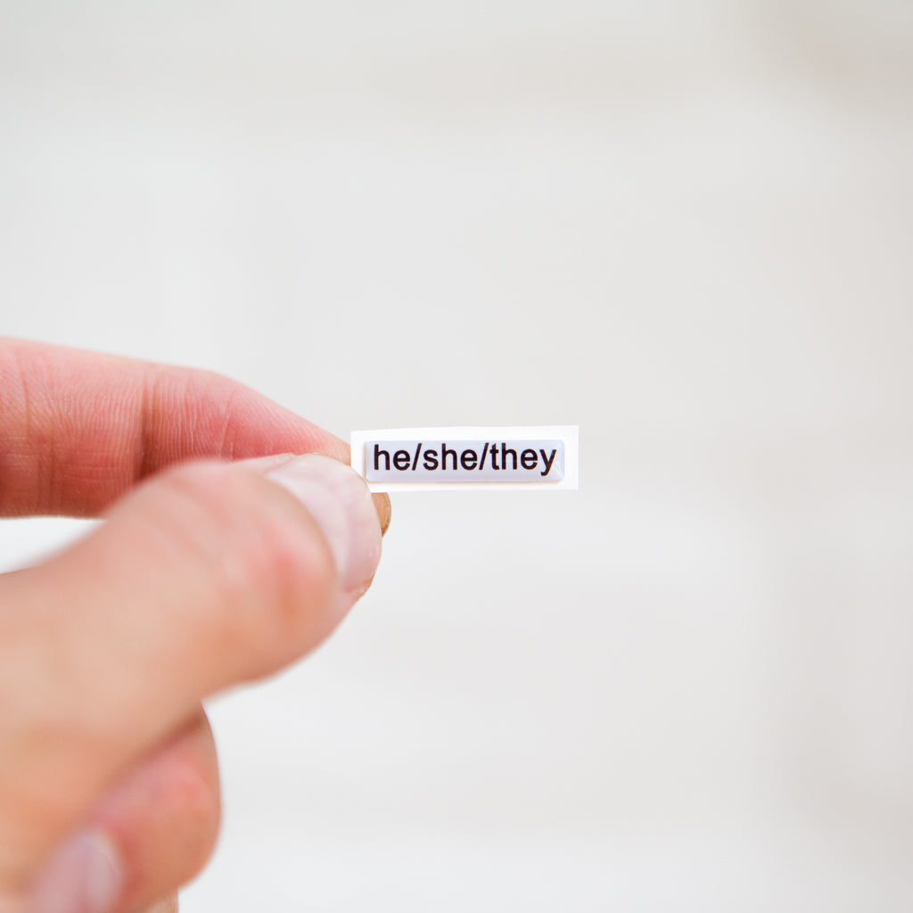 A white sticker with black text displaying he/she/they pronouns, designed for name badges and ID tags. A high-quality pronoun sticker with durable epoxy resin. This pronouns sticker for name badges