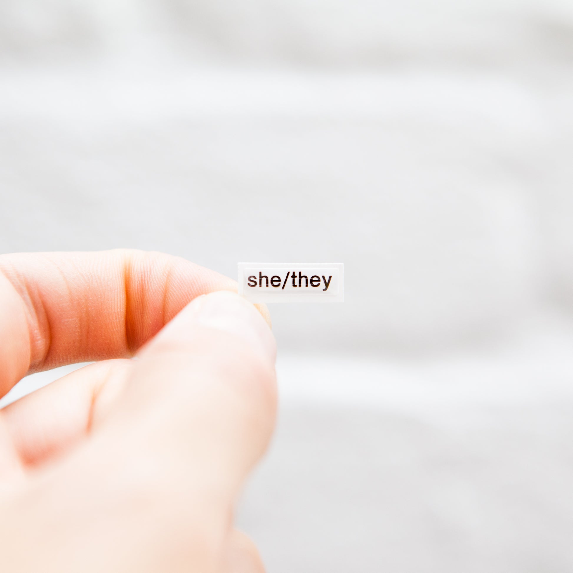 A white sticker with black text displaying she/they pronouns, designed for name badges and ID tags. A high-quality pronoun sticker with durable epoxy resin. This pronouns sticker for name badges