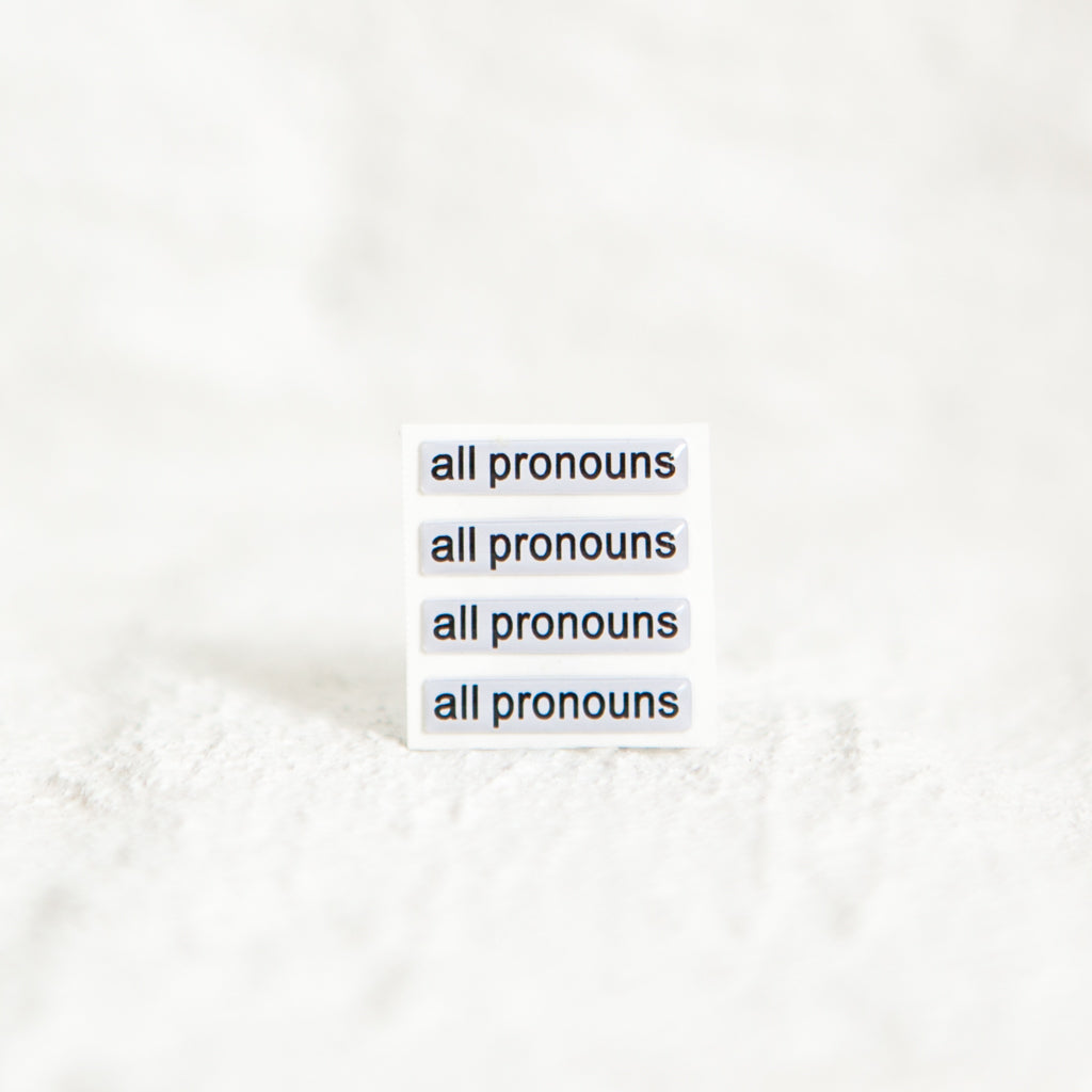 A white sticker with black text displaying 'all pronouns', designed for name badges and ID tags. A high-quality pronoun sticker with durable epoxy resin. This pronouns sticker for name badges