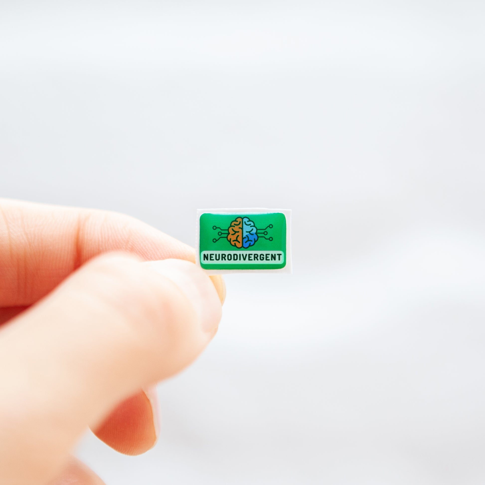 Fingers holding a green sticker, with 'Neurodivergent' text, and a digitally illustrated image of a brain, representing neurodivergent stickers.